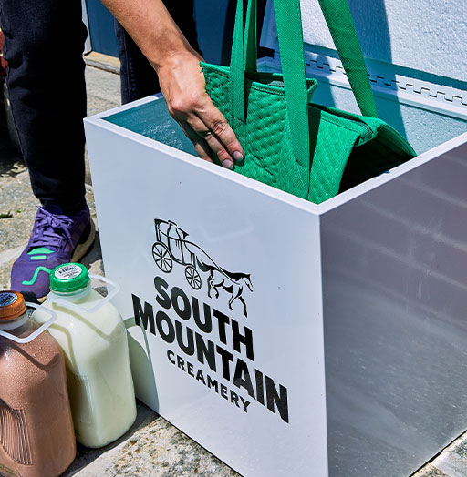 Photo of a green SMC cooler bag being lowered into an SMC porch box. Next to the porch box are two gallons of SMC milk