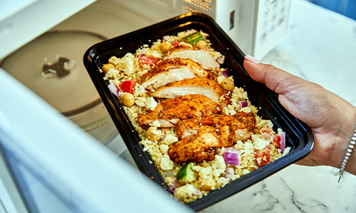 Person removes an SMC home delivery meal from the microwave; the dish includes quinoa and grilled chicken